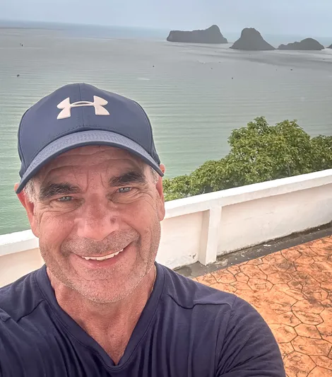 David Hibbins – Author Portrait at Coastal Viewpoint in Thailand David Hibbins smiling in a navy cap with ocean cliffs in the background in Thailand