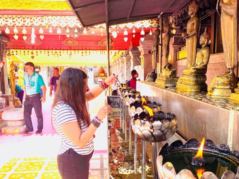 Woman lighting a candle at a Buddhist temple in Thailand, with Buddha statues and rows of flames beside her.