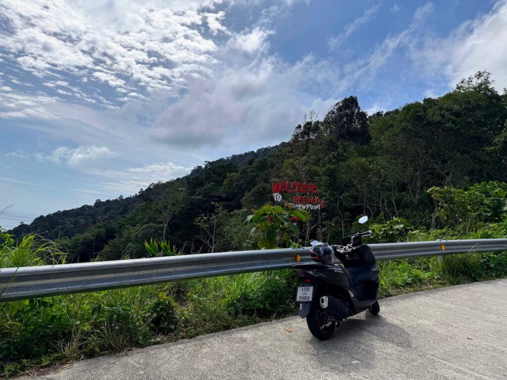 Scooter parked beside a mountain road at a viewpoint in Thailand, surrounded by lush green hills under a dramatic sky.