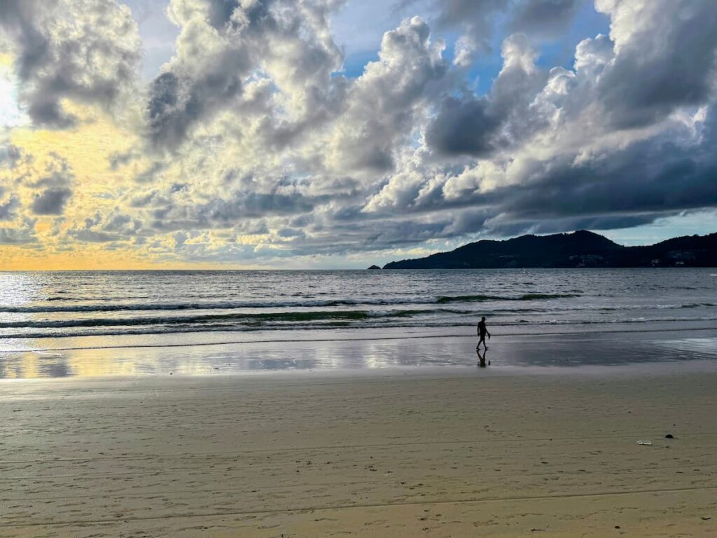 Dramatic clouds over Patong Beach in Phuket with a lone person walking along the shoreline.